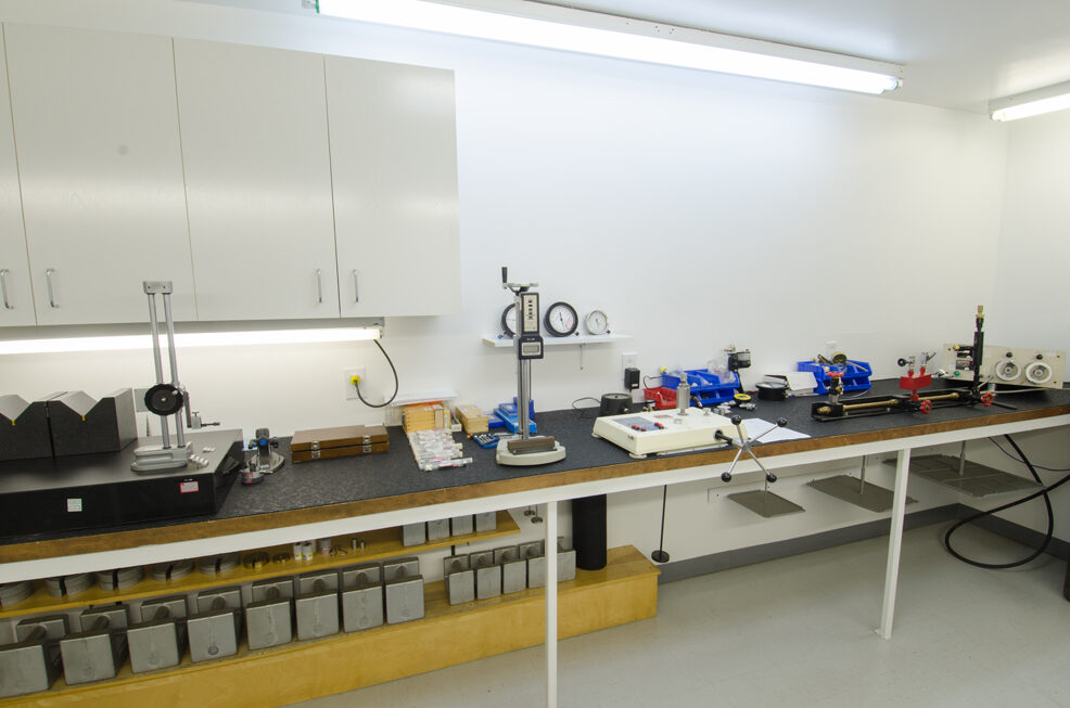 Etaltech’s calibration laboratory: a workbench with measuring instruments and dial indicators on stands, and calibration weights stored on the lower shelf